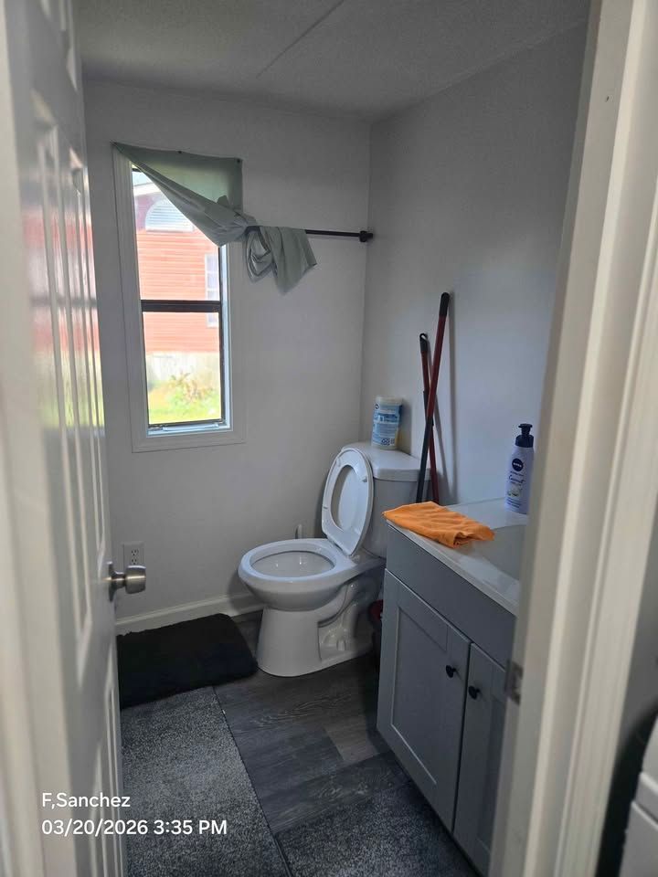 Small bathroom with toilet, gray cabinet, cleaning tools, and a window with a green curtain.