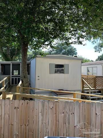 Small beige mobile home behind a wooden fence and deck, shaded by a large tree in a trailer park.