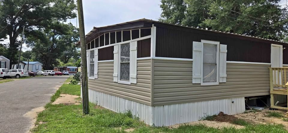 Small beige-and-brown mobile home with white shutters beside a gravel road and trees