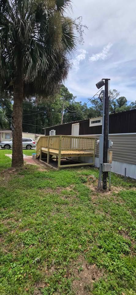 Wooden ramp leading to a mobile home, with a palm tree and grassy yard beside a metal post