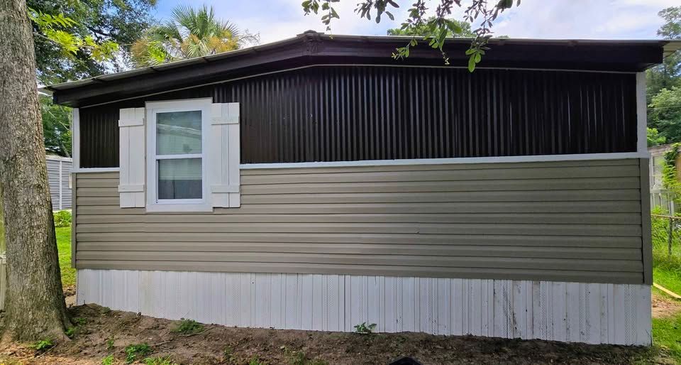 Small gray house with white trim, dark upper siding, and a window, surrounded by trees.