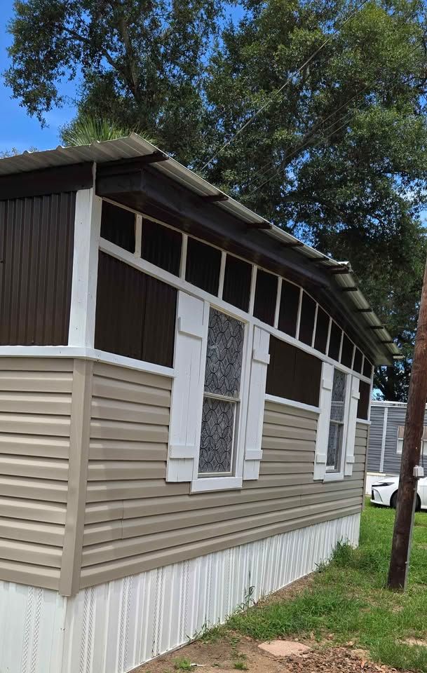Side view of a beige and brown mobile home with white trim and screened windows under trees.