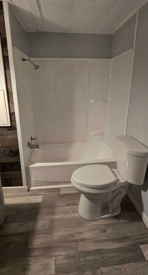 Small bathroom with white tub-shower combo, toilet, and gray wood-look flooring