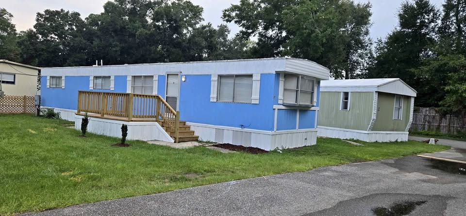 Light blue mobile home with a wooden ramp in a grassy yard, with another small white trailer nearby.