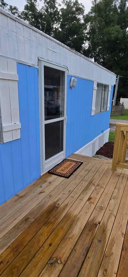 Blue tiny house with a white-trim door and wooden deck surrounded by trees