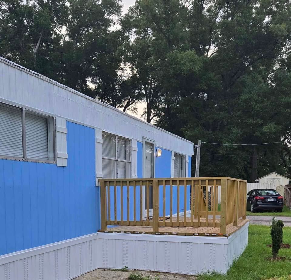 Blue mobile home with a small wooden porch, trees behind it, and a car parked nearby.