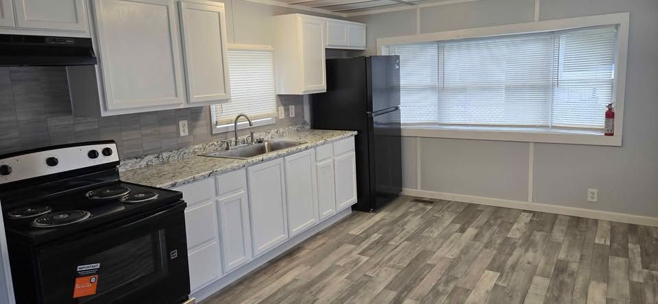 Empty kitchen with black appliances, white cabinets, and a large window over wood-look flooring.