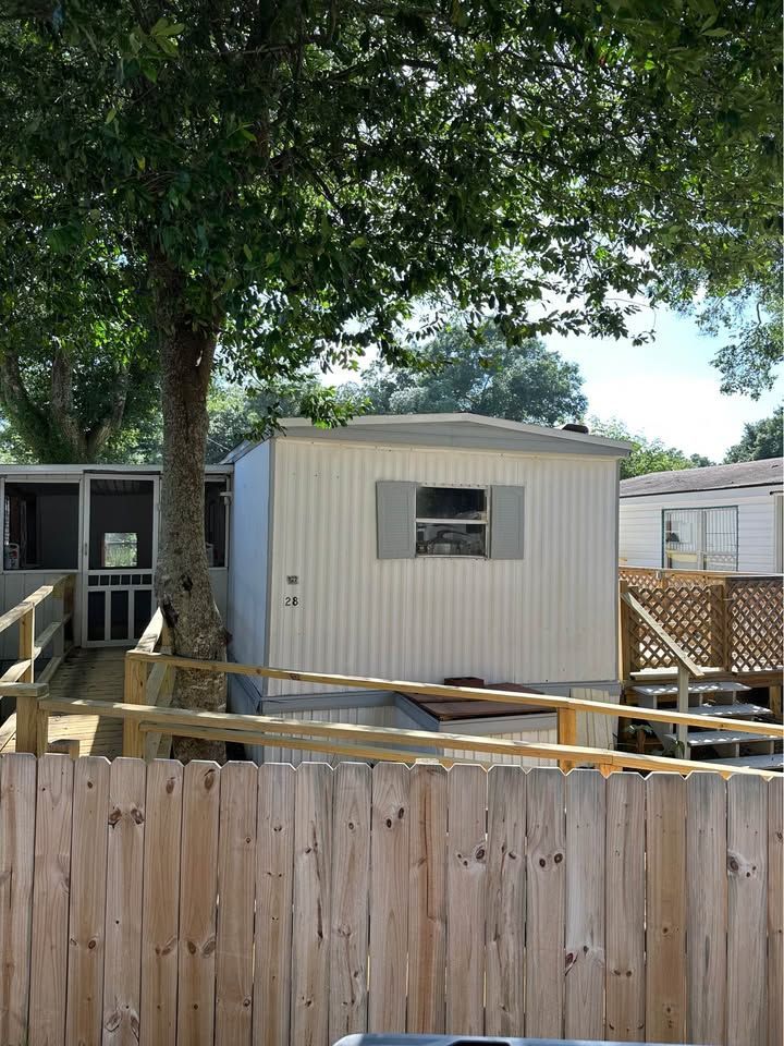 A beige mobile home in a fenced yard with a large tree overhead and a wooden deck in front