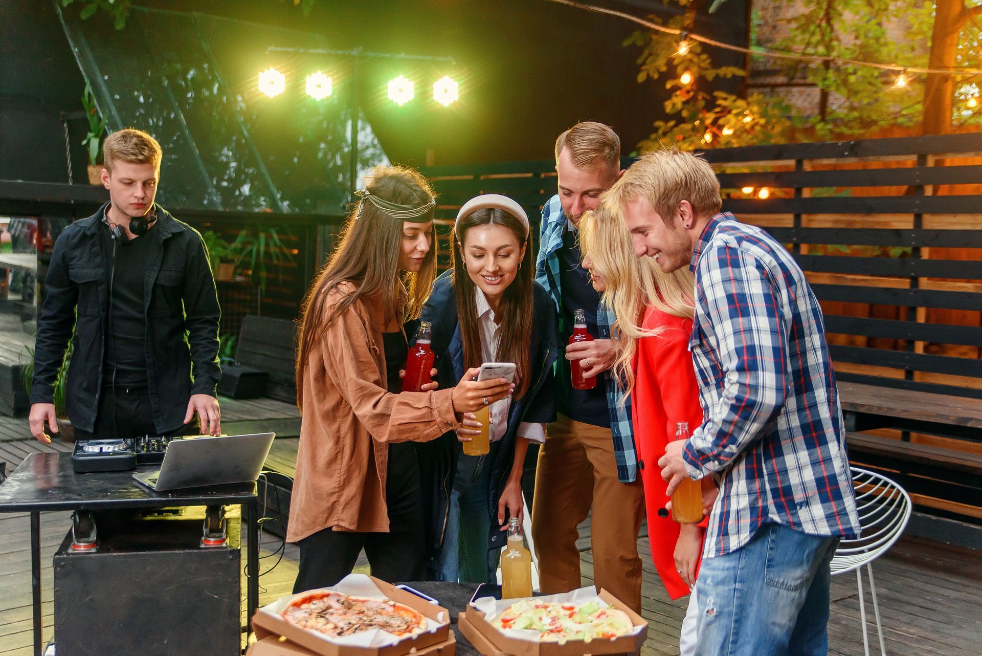 Group of friends looking at a phone, pizza boxes on a table, DJ in the background, outdoor party.