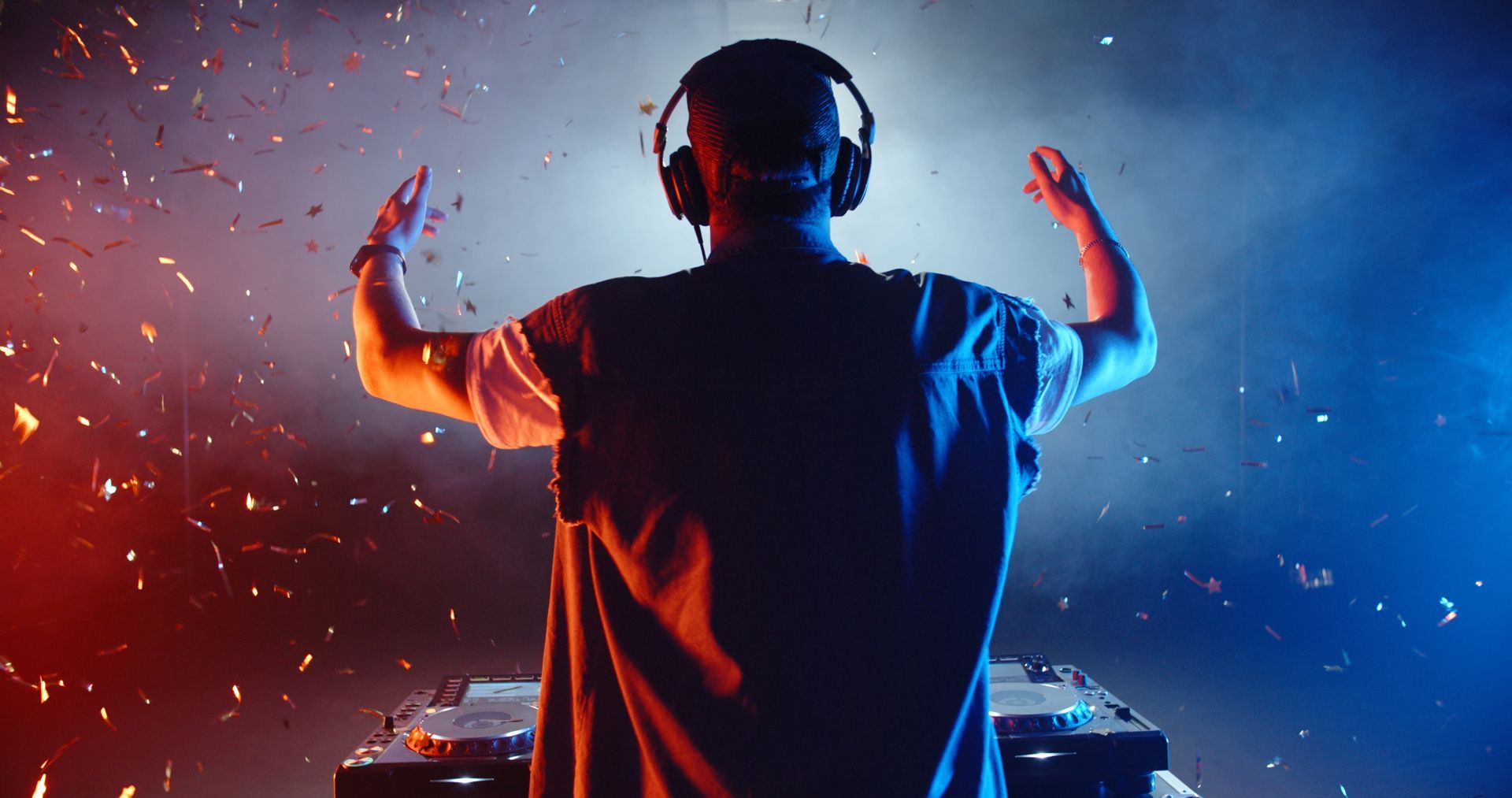 DJ with headphones raising hands in a smoky, colorful environment behind a soundboard.
