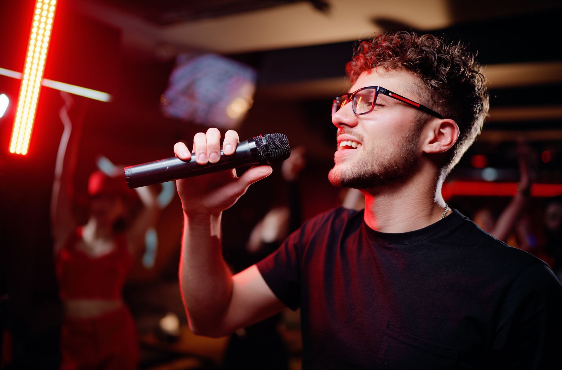 Man singing into a microphone in a dimly lit venue with red lights.