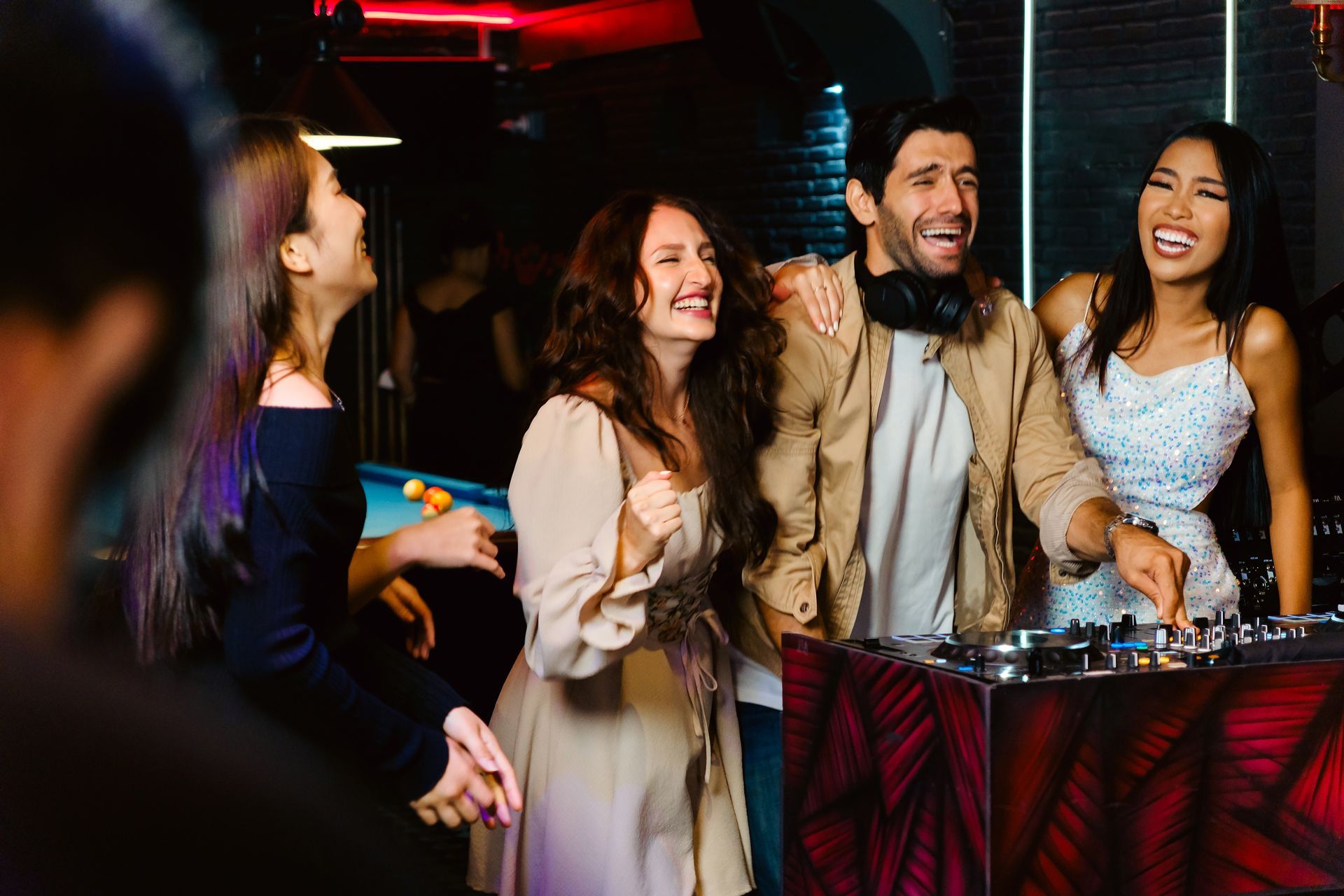 People laughing and dancing at a nightclub, next to a DJ set and a pool table.