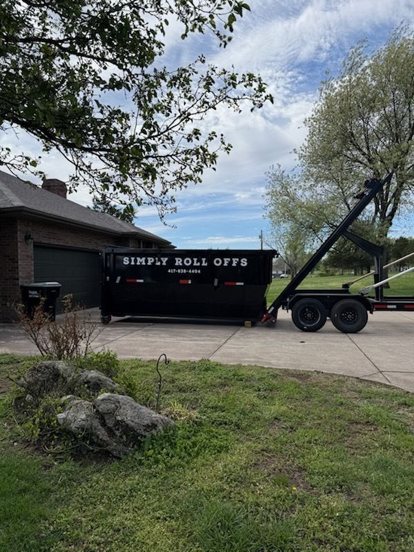 Black dumpster trailer parked in a driveway beside a house and garage, with trees and grass around it