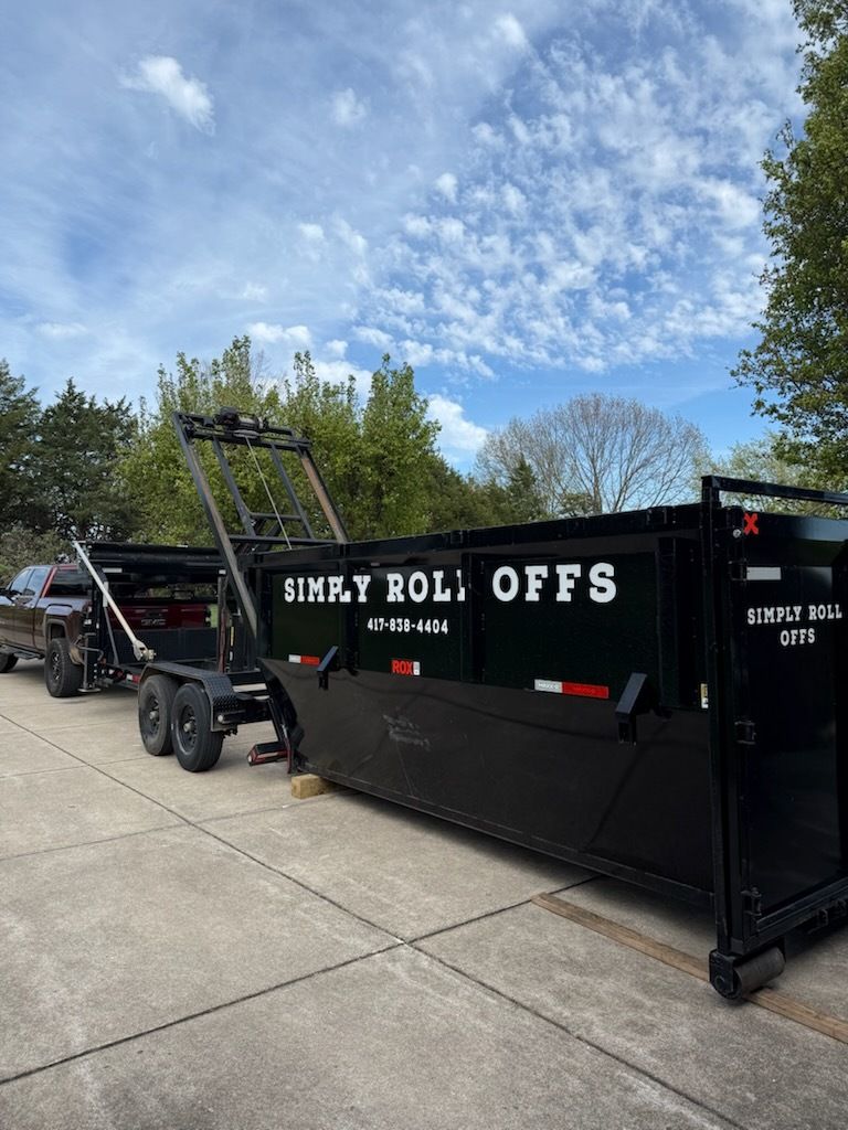 Black dumpster trailer parked on a driveway under a blue sky, with white lettering and a hoist frame.