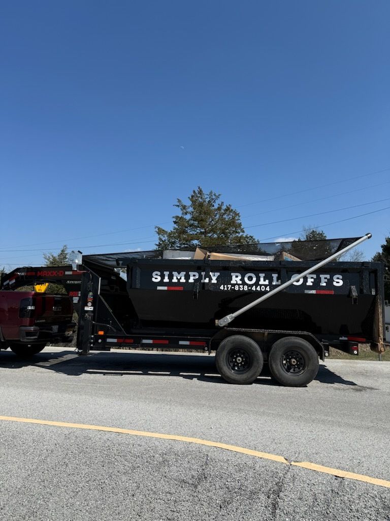 Black dump trailer with “SIMPLY ROLL OFFS” parked on a sunny road under a clear blue sky.