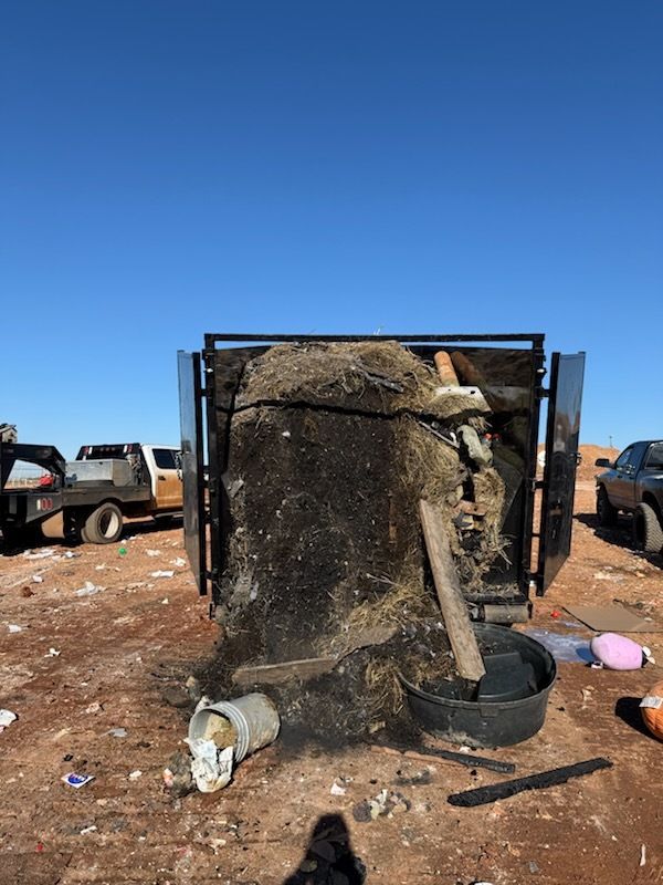 Overturned black water tank in a dirt lot, with debris and a truck under a clear blue sky.