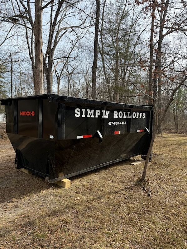 Black dumpster labeled “SIMPLY ROLL-OFFS” in a wooded area with bare trees and dry ground