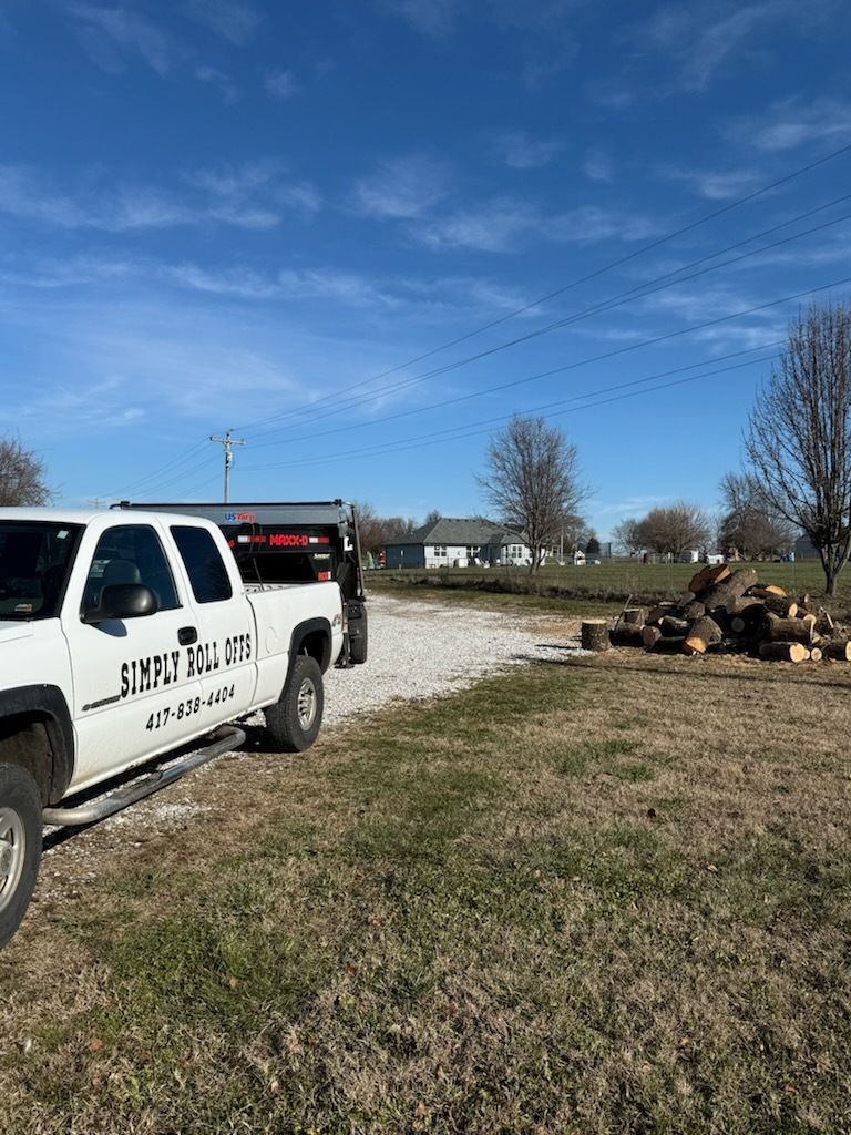 White work truck beside a dirt road near a pile of cut logs under a clear blue sky