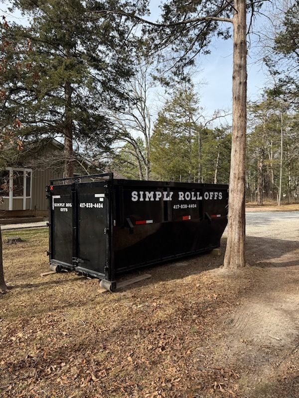 Black Simple Roll-Offs dumpster in a wooded yard beside a house and trees