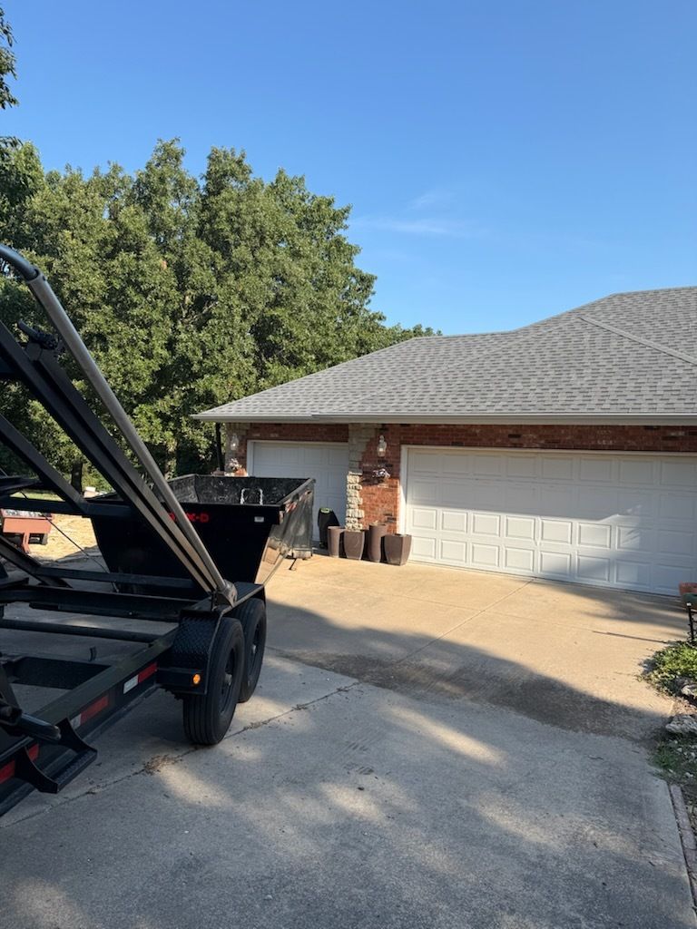 Driveway beside a brick house with a white garage, trees in back, and a black trailer in the foreground