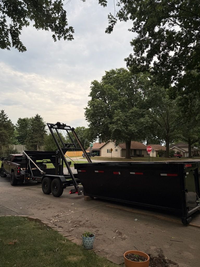 Black dump trailer being loaded by a skid steer on a tree-lined street with houses in the background