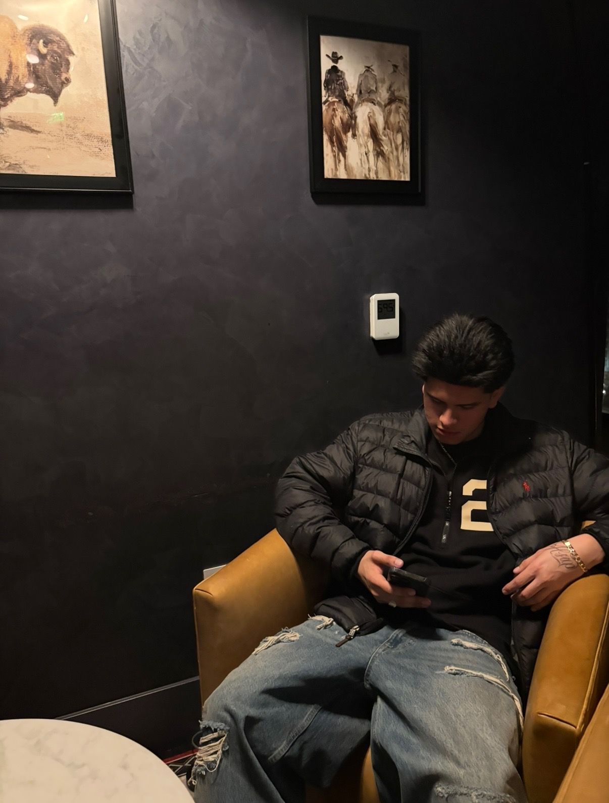 Person sitting in a tan chair, looking at a phone in a dark room with framed wall art.