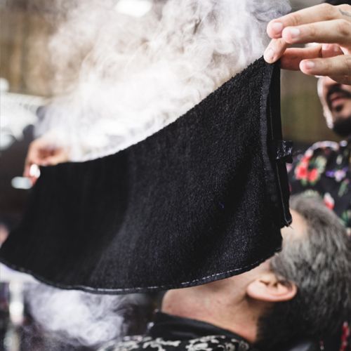 Barber steaming client's face with hot towel; black towel, steam rising, close up.