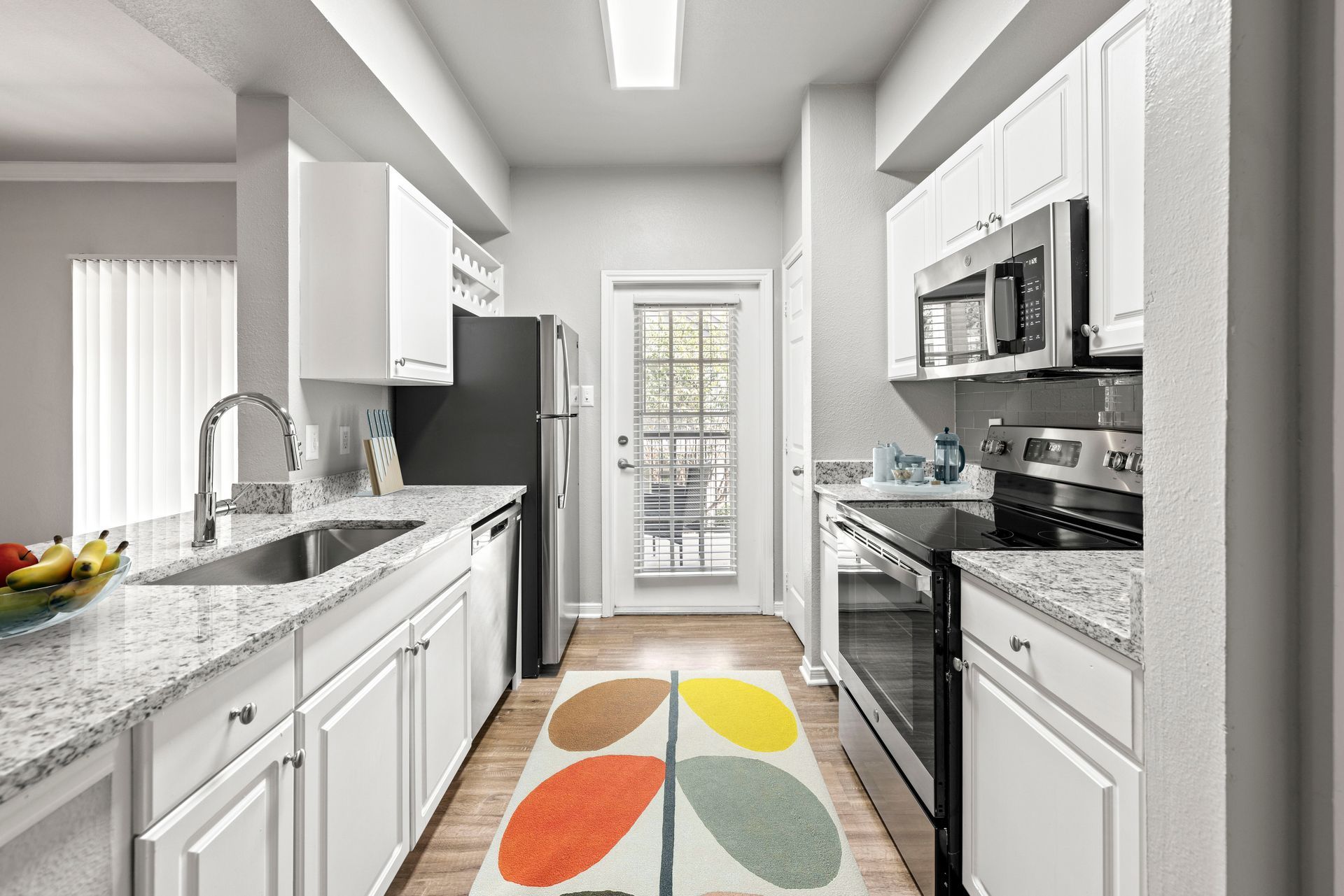 Bright galley kitchen with white cabinets, granite counters, stainless appliances, and a colorful runner rug.