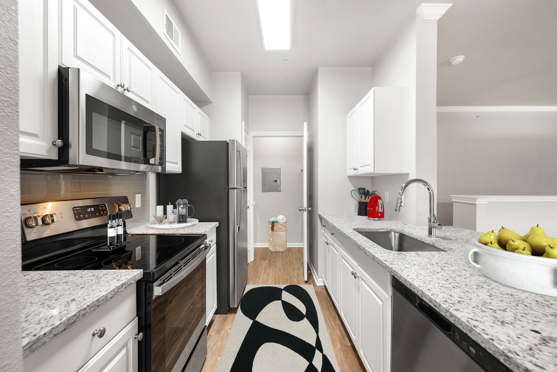 Bright narrow kitchen with stainless appliances, white cabinets, gray counters, and a bowl of fruit on the island.