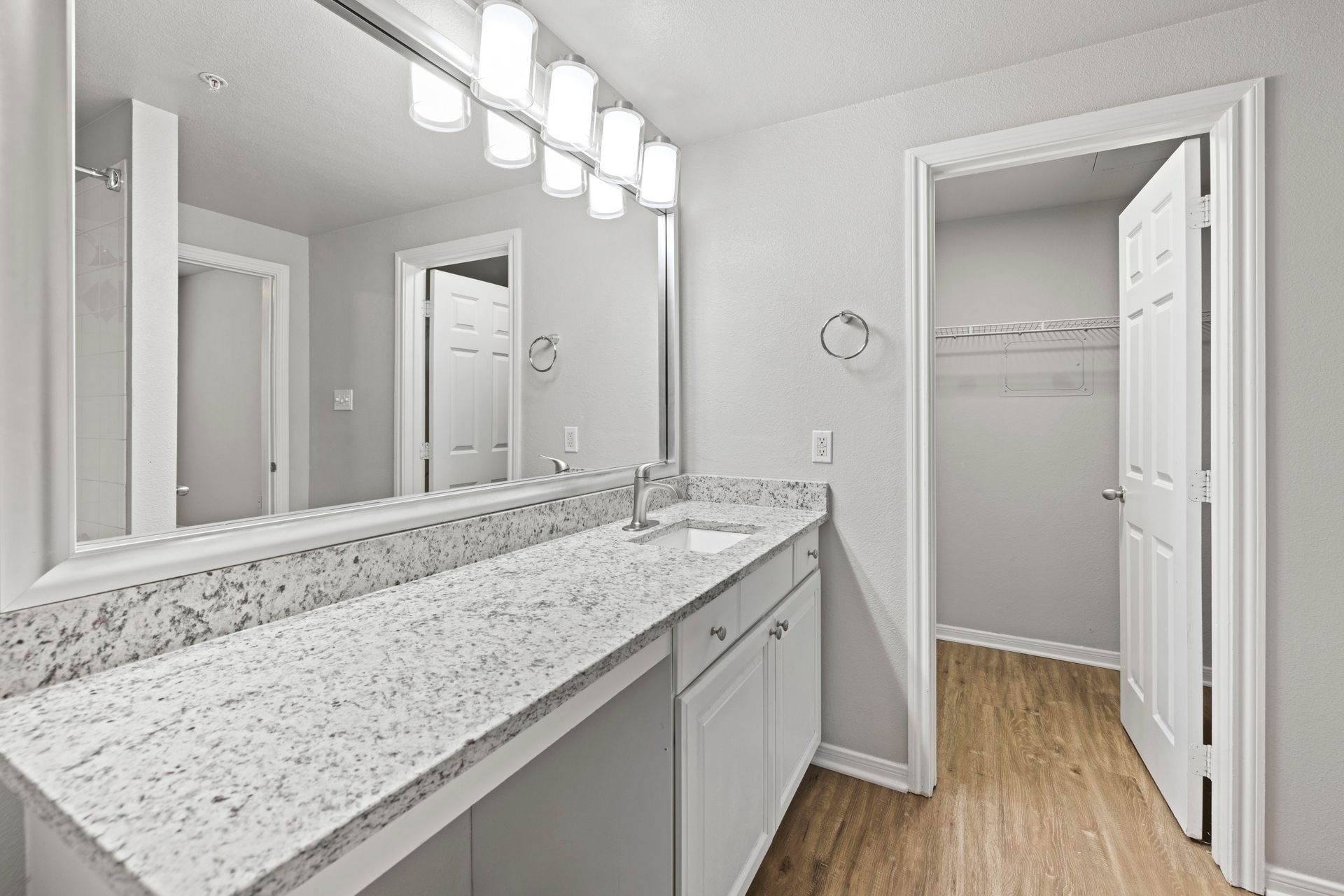 Bright bathroom with double vanity, large mirror, and open closet doorway adjacent to wood flooring.
