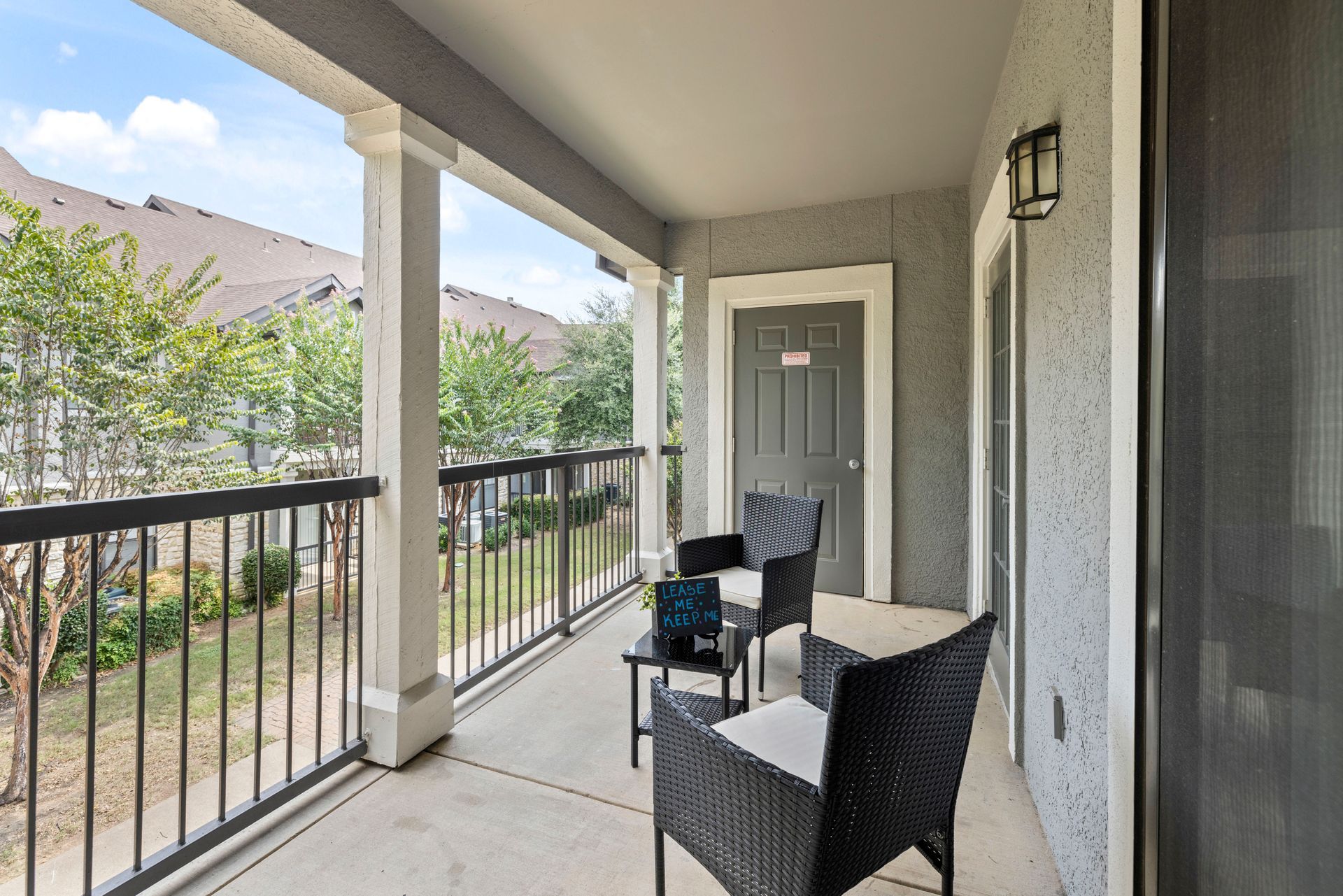 Covered apartment balcony with two chairs, black railing, and gray front door overlooking trees and neighboring homes