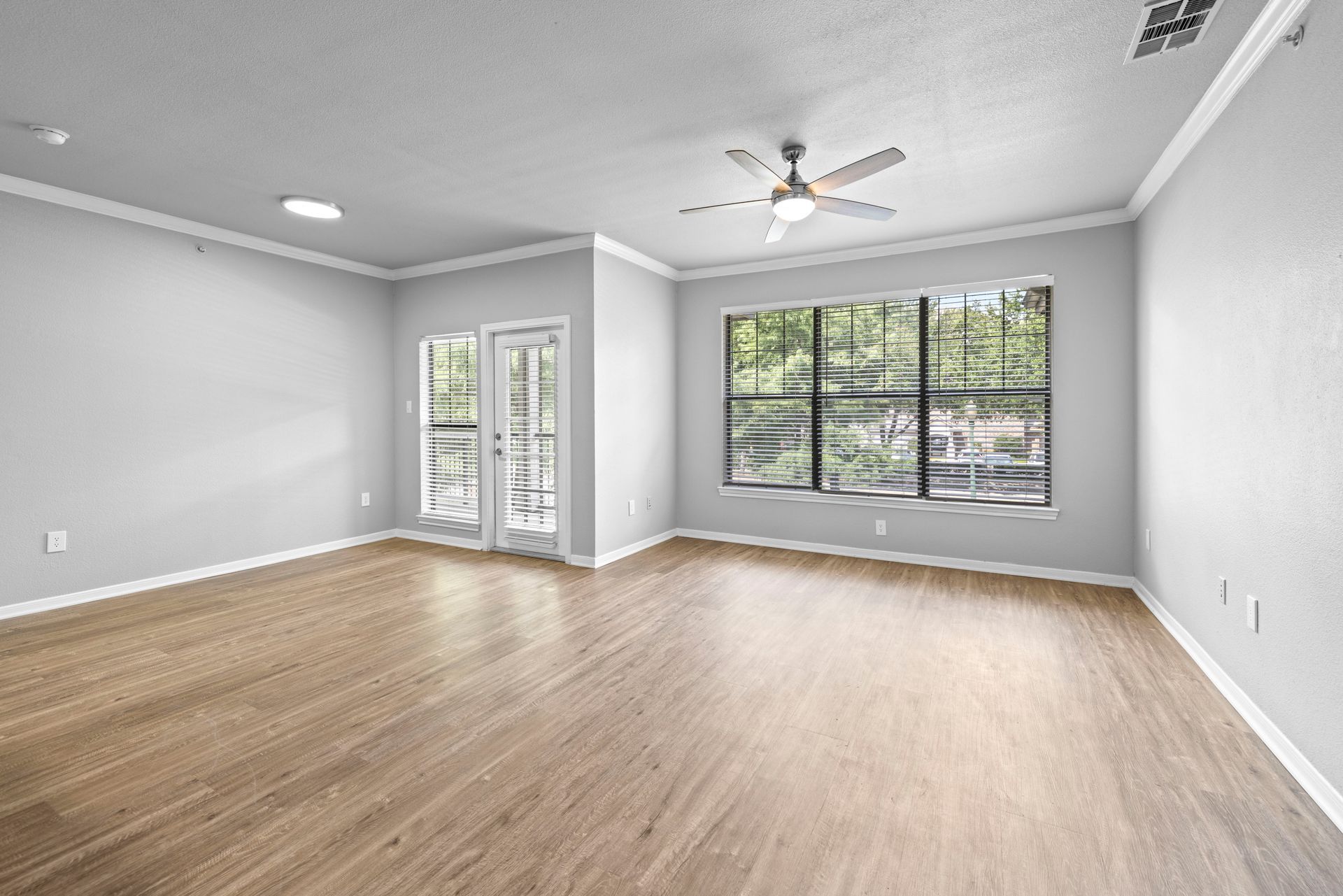 Empty modern living room with gray walls, wood flooring, ceiling fan, and large windows.