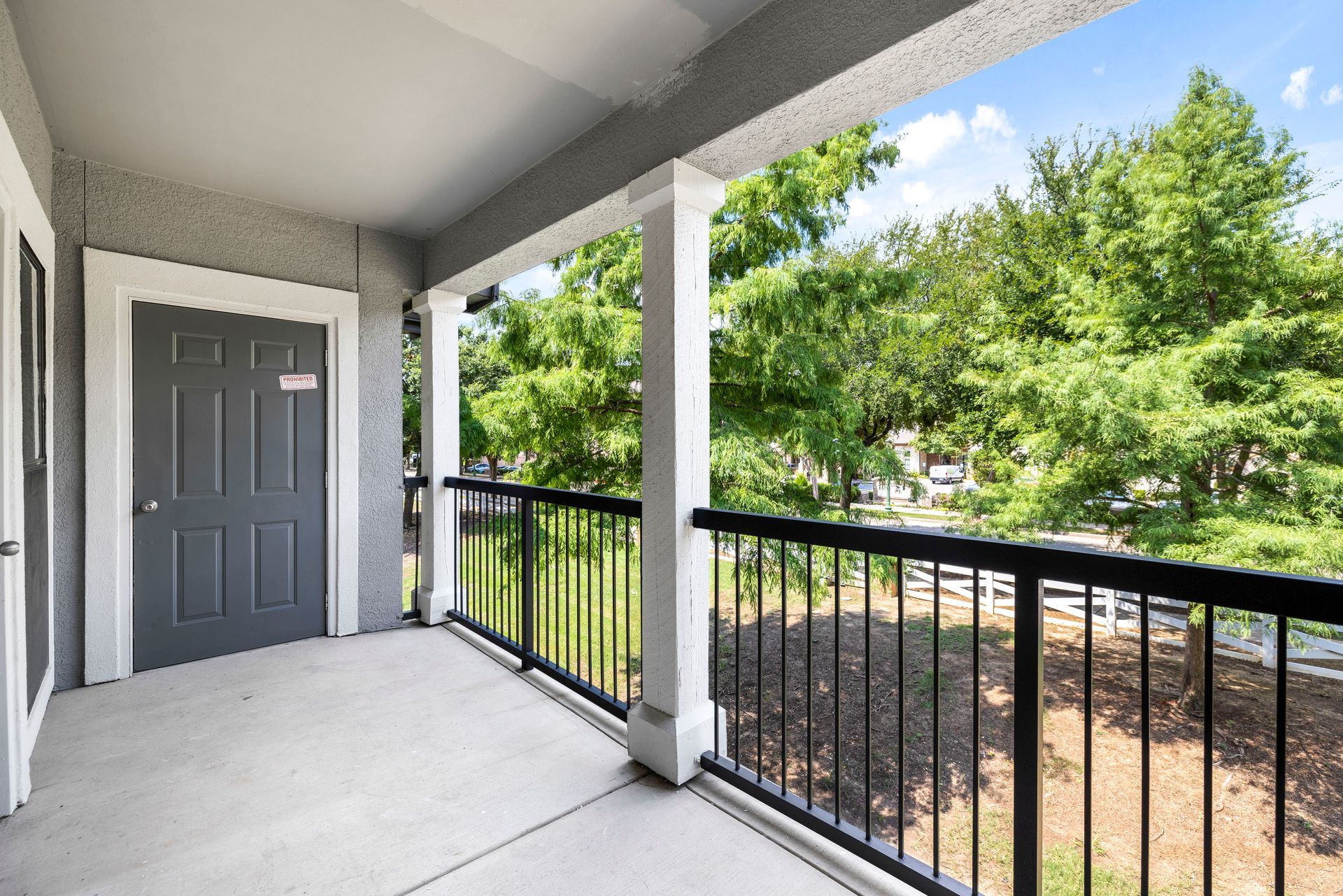 Covered apartment balcony with gray door, black railing, and trees outside