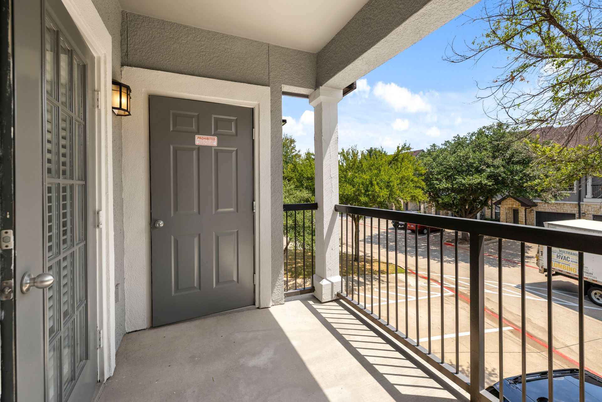 Apartment balcony with gray door, black railing, and view of trees and parking lot