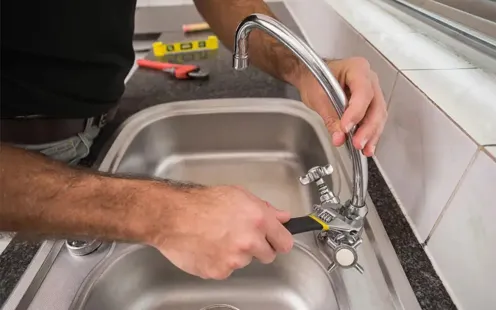 GTZ Plumbing Service LLC - A man is fixing a faucet in a kitchen sink with a wrench.