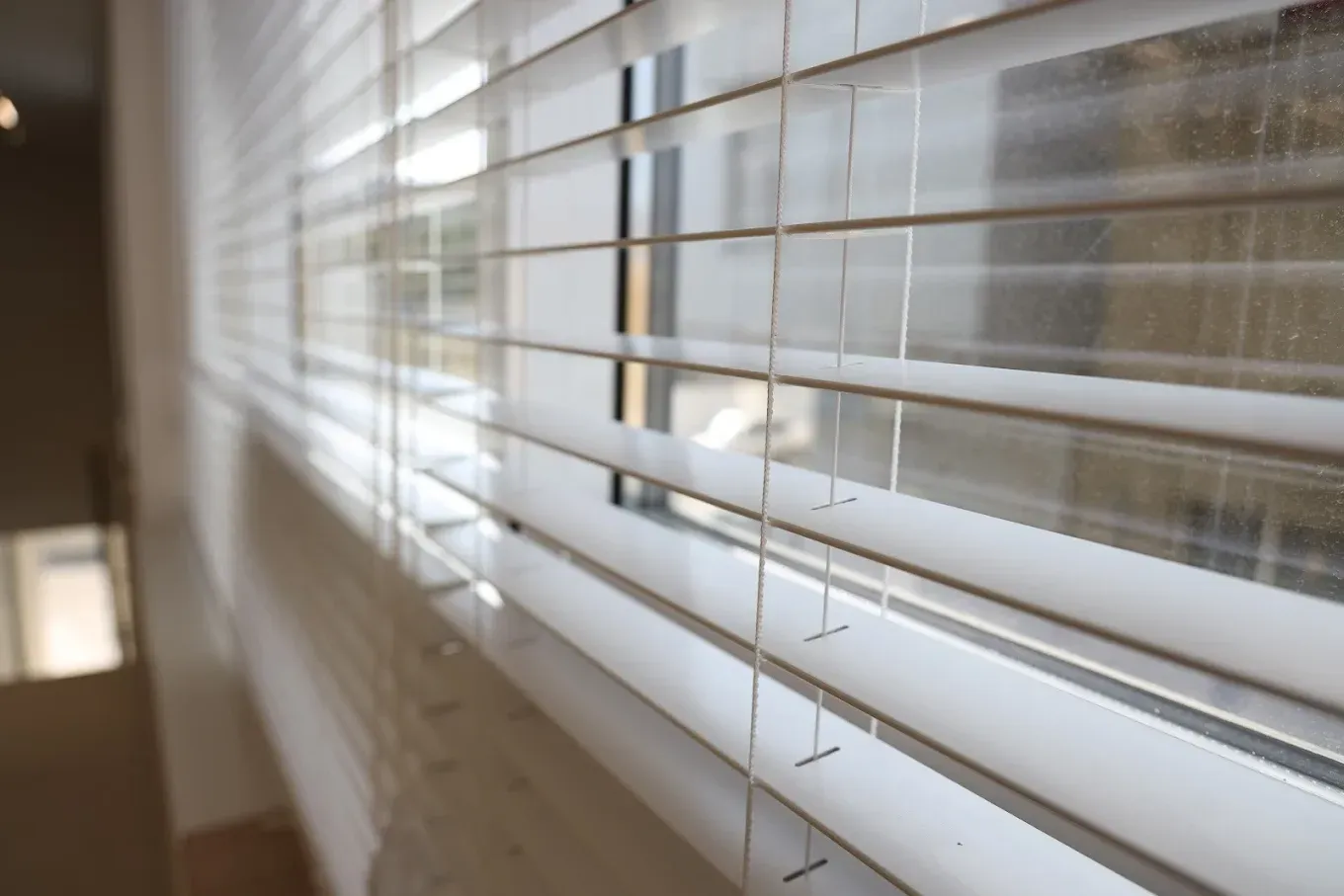White horizontal window blinds partially closed, allowing light to shine through from a window.