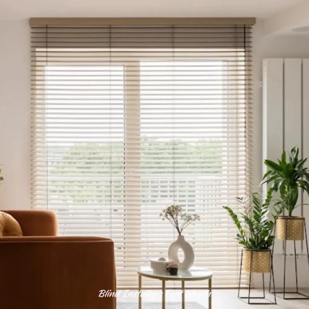 A bright room with a large window covered by light wood venetian blinds, featuring a velvet armchair and potted plants.