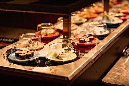 Sushi dishes on a conveyor belt in a restaurant. Various plates with covered sushi rolls are visible.
