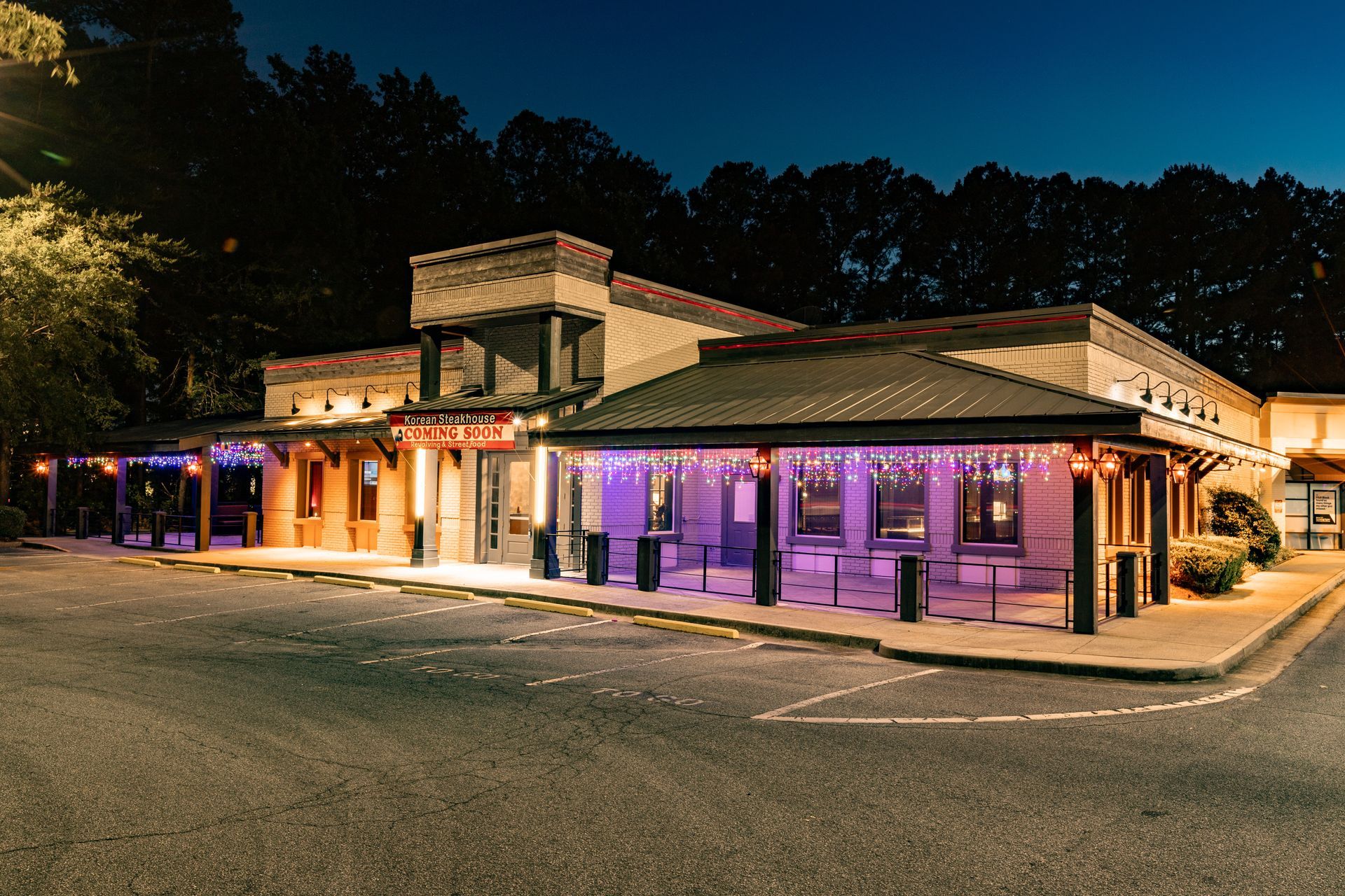 Nighttime exterior of a restaurant lit with colorful lights, featuring an outdoor seating area. The building is a tan color, with trees in the background.