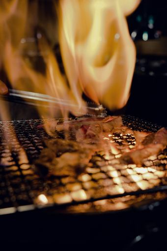 Meat grilling over an open flame on a metal grate. Tongs hold the meat, and large yellow flames rise up.