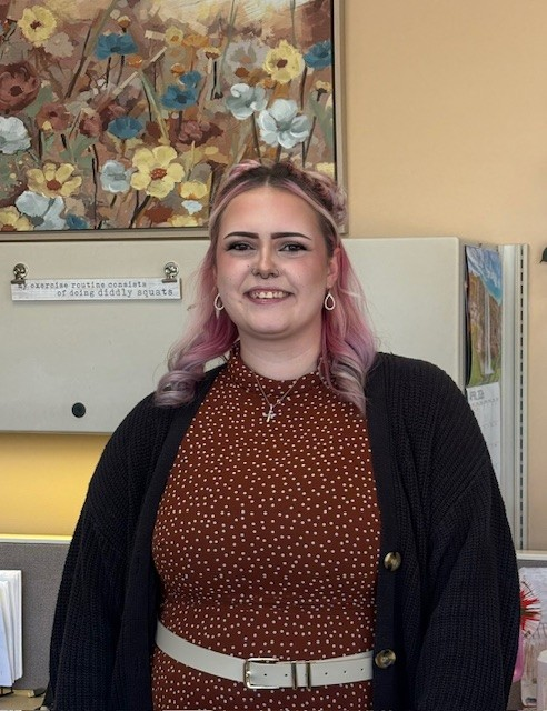 Woman standing in an office, smiling, with pink hair, burgundy dress, and dark cardigan. Woman standing in an office, smiling, with pink hair, burgundy dress, and dark cardigan.