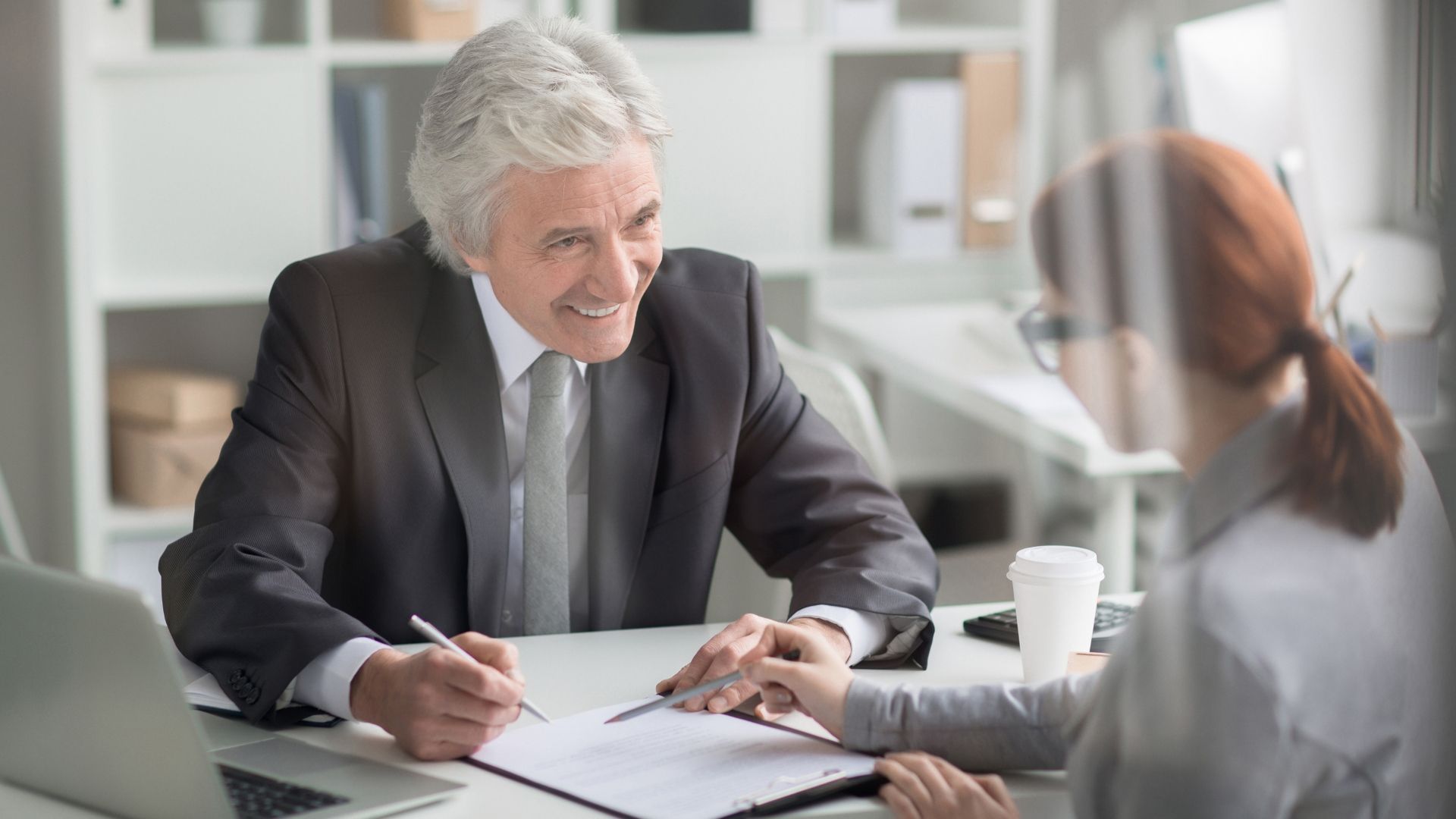 Older man in a suit signs a document, smiling at a woman; office setting.