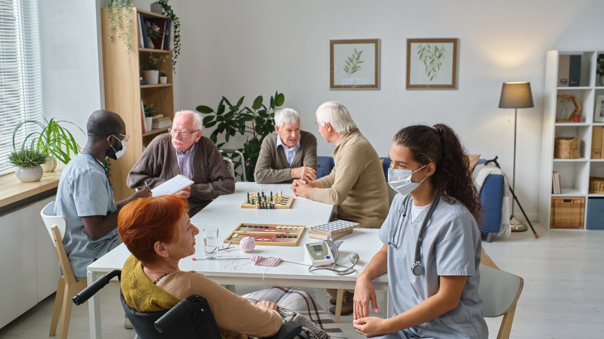 Group of people in a room, including elderly and healthcare workers, gathered around a table with games.