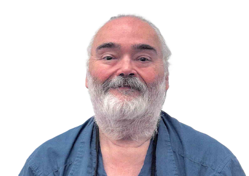 Man with a white beard and graying hair, wearing blue scrubs, smiling at the camera.