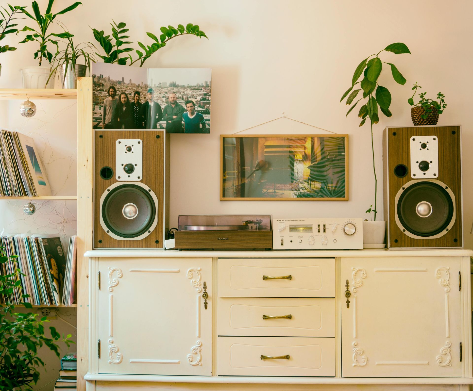 Record player and speakers on a white cabinet with records, plants, and art in a cozy setting.