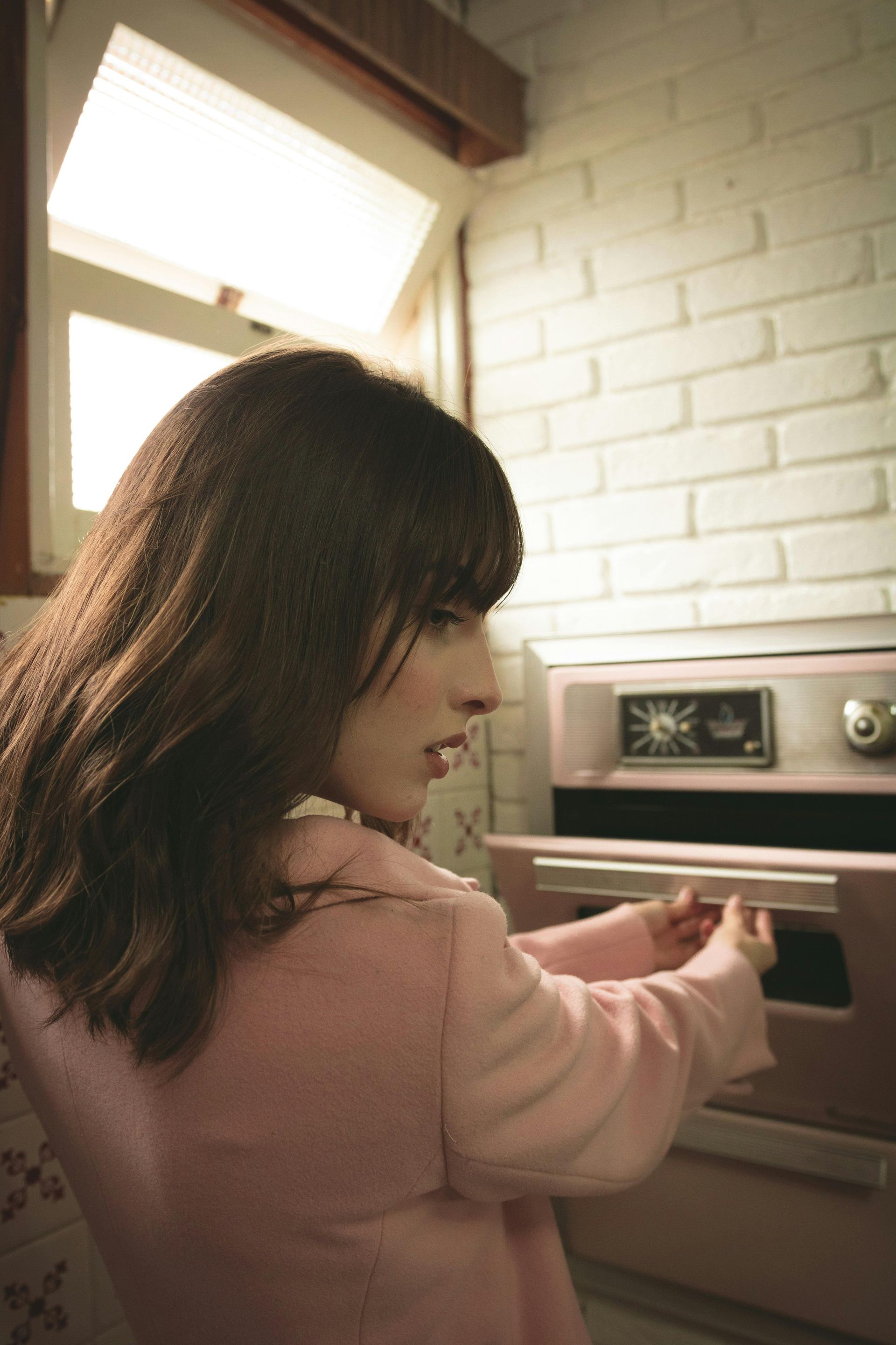 Woman in pink jacket opening a pink oven in a kitchen with white brick walls.