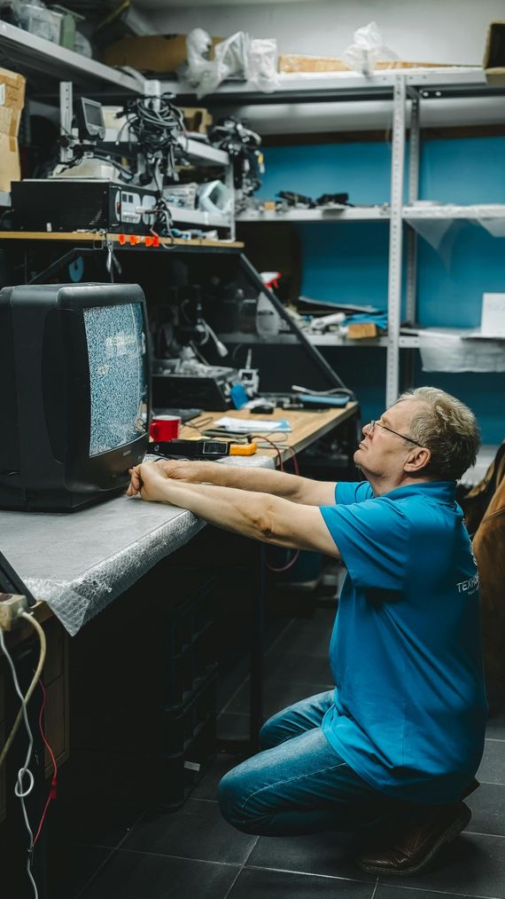 Man kneels before a static-filled TV in a cluttered workshop. He leans forward, focused.