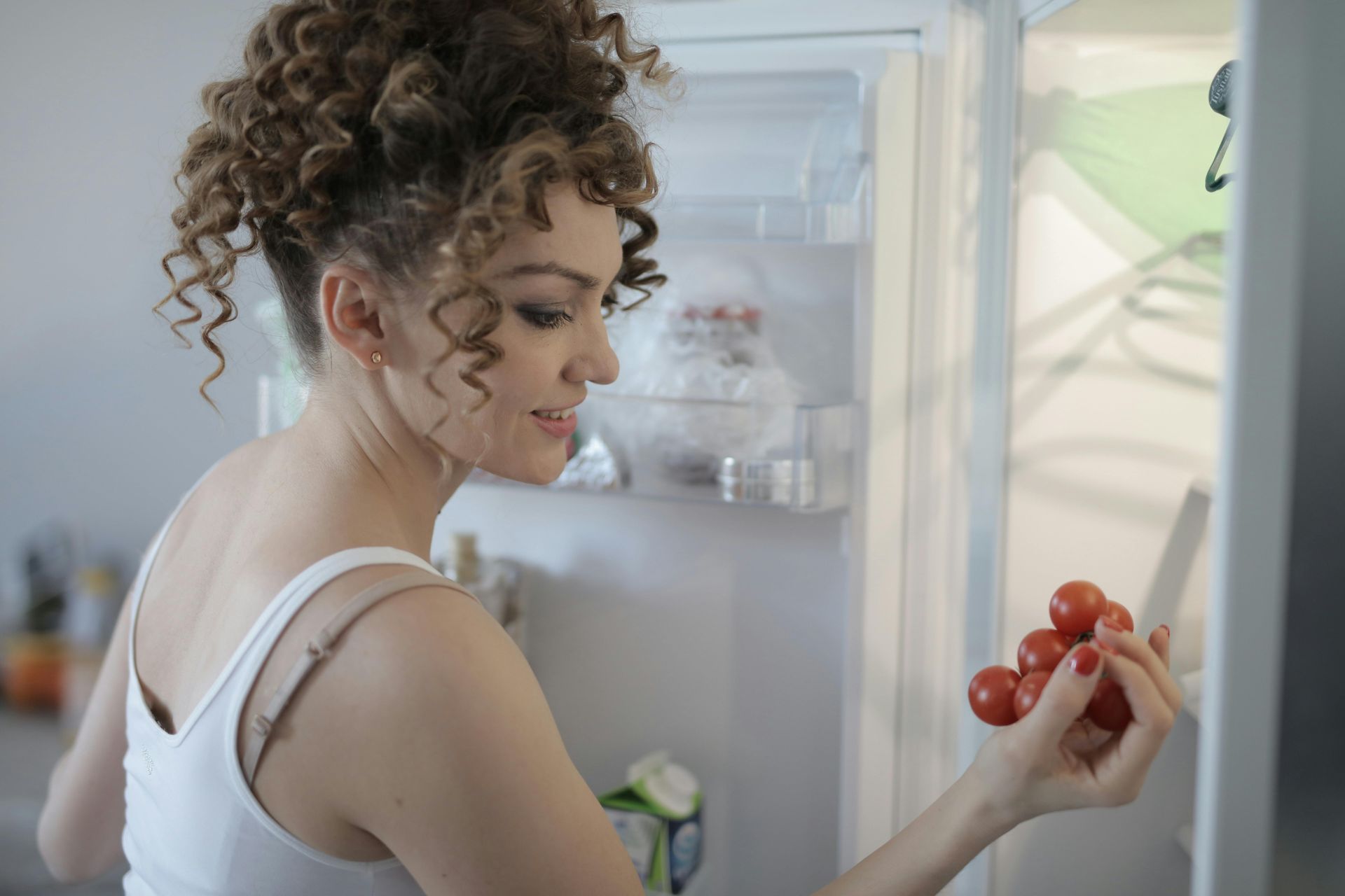 Woman in a white tank top looks at tomatoes while standing in front of an open refrigerator.