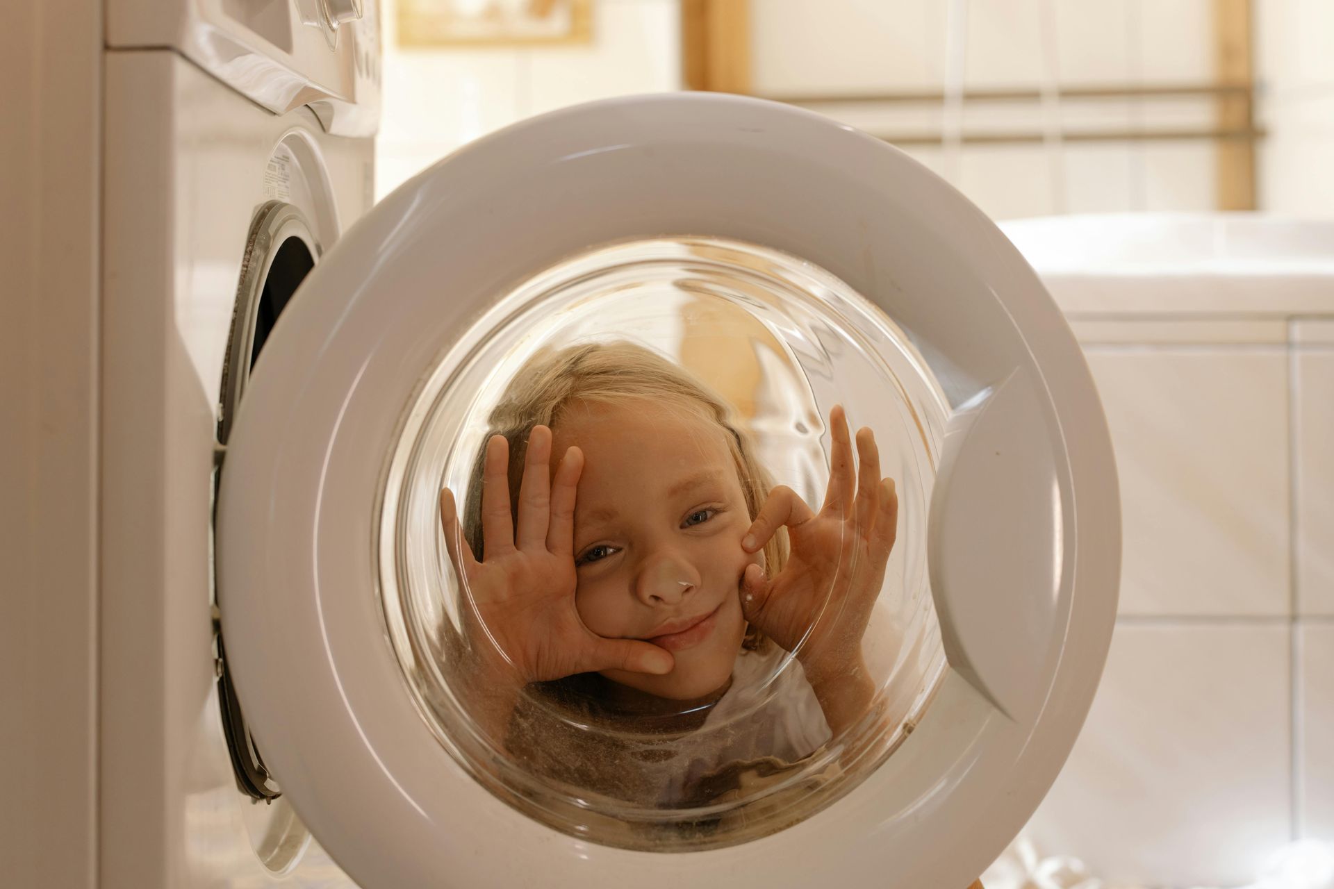 Child smiling, inside an open washing machine, hands against the glass.
