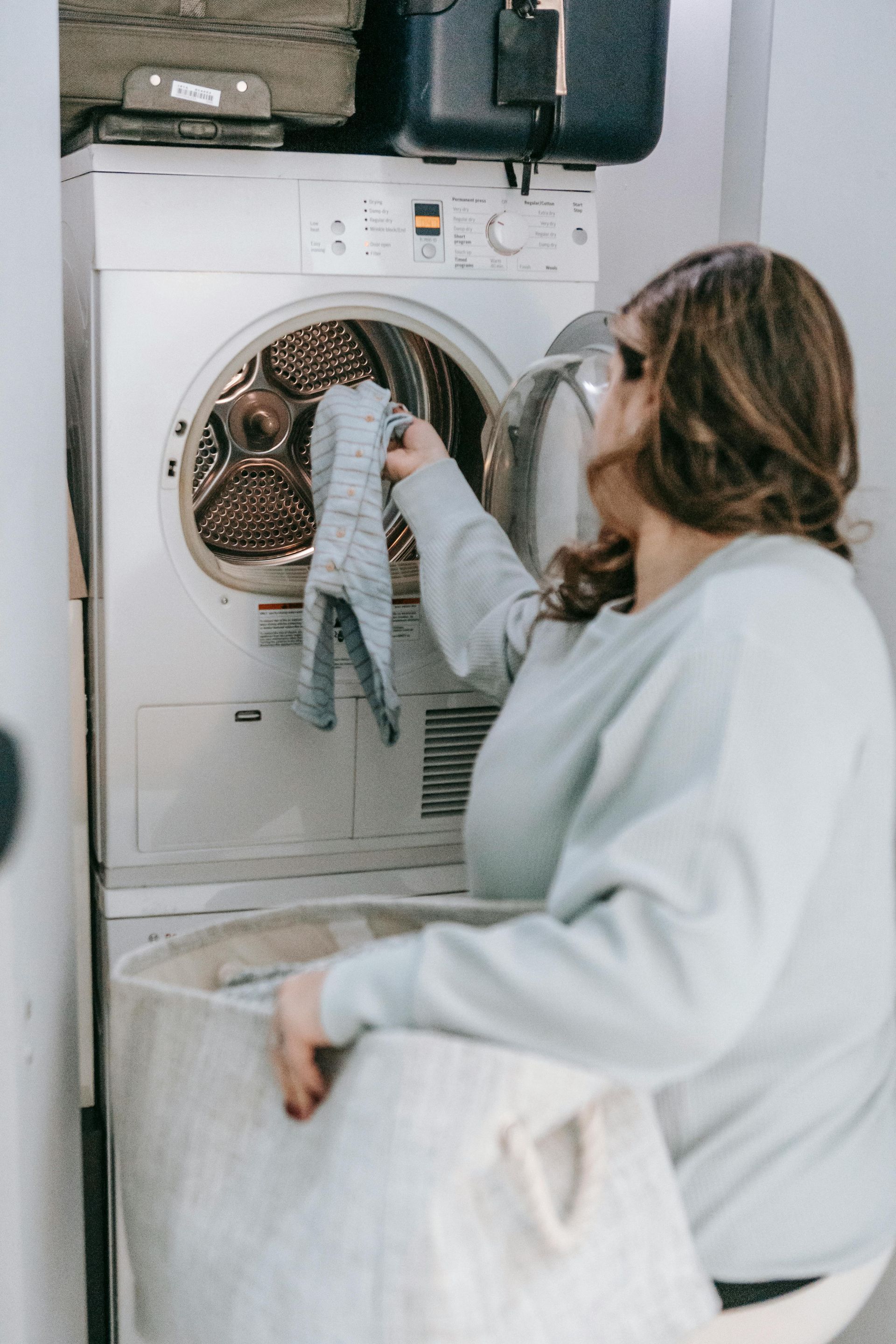 Woman removing clothes from dryer, holding a gray item. Laundry basket in front.